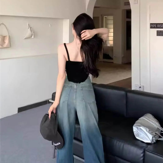 Woman in a black tank top and wide-leg jeans holding a black cap, standing in a modern living room.