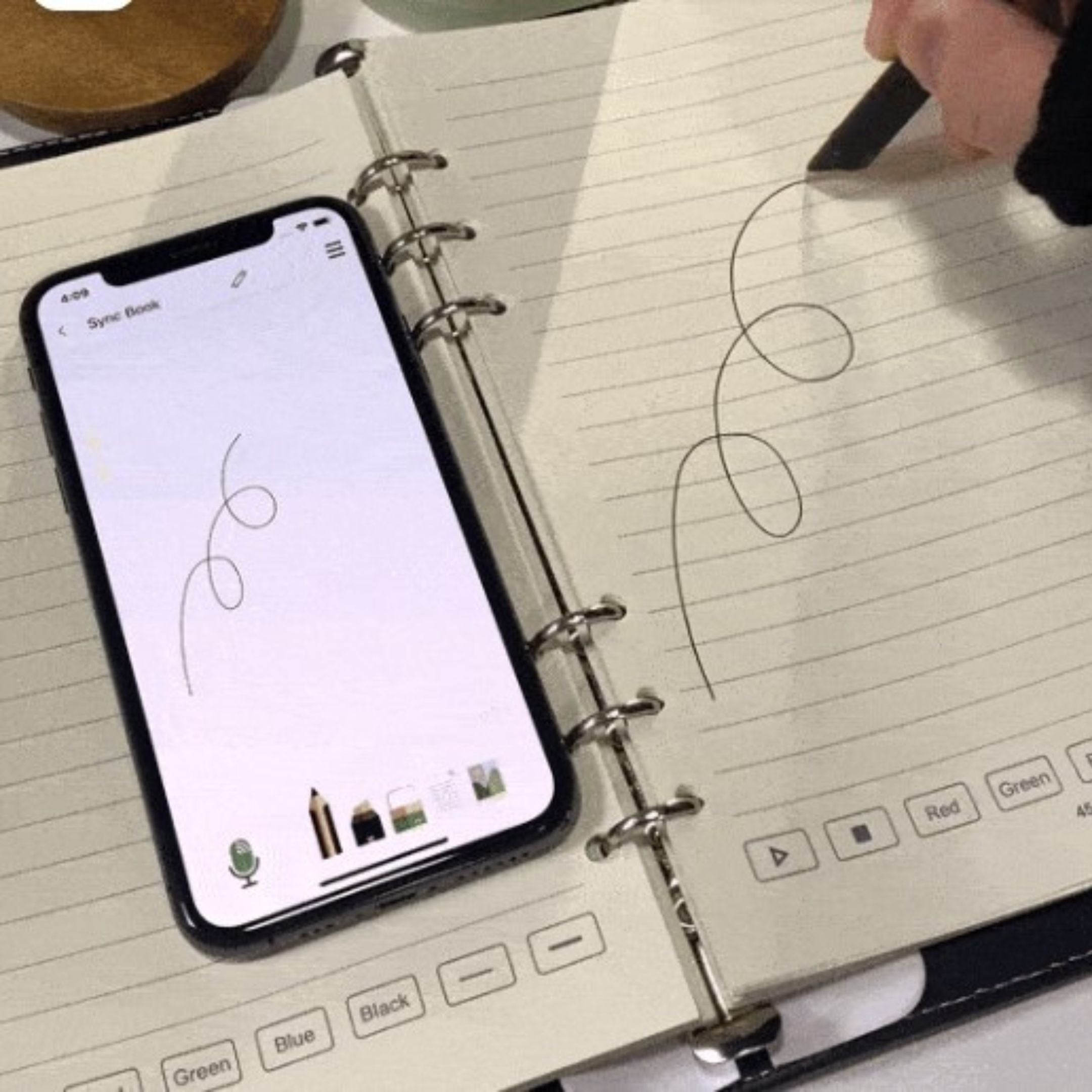 A smartphone with a scribble displayed on its screen is placed on an open notebook with a similar scribble. A hand holding a pen is drawing the same pattern on the notebook. The notebook has lined pages.