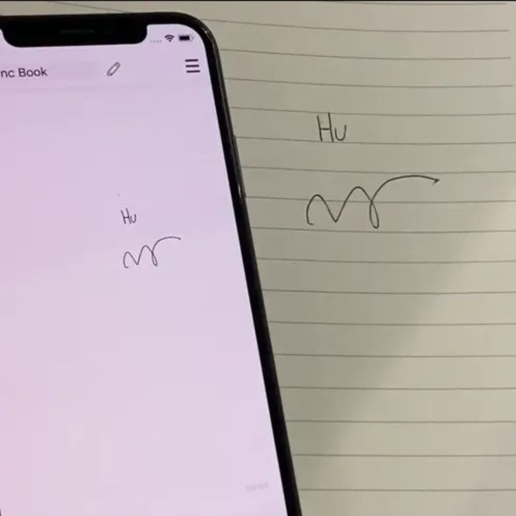 A smartphone displaying handwritten text and a wavy line, which are mirrored on a lined notebook beside it. The word Hu is visible on both the phone screen and paper.