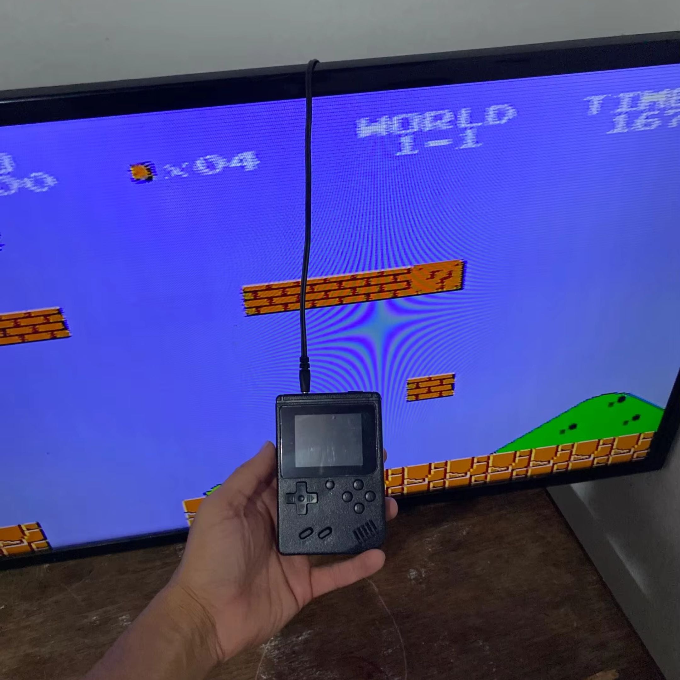 A person holds a small, black handheld gaming device connected to a large screen displaying a pixelated video game with bricks, sky, and a green hill. The game has text showing WORLD 1-1 and various numbers.