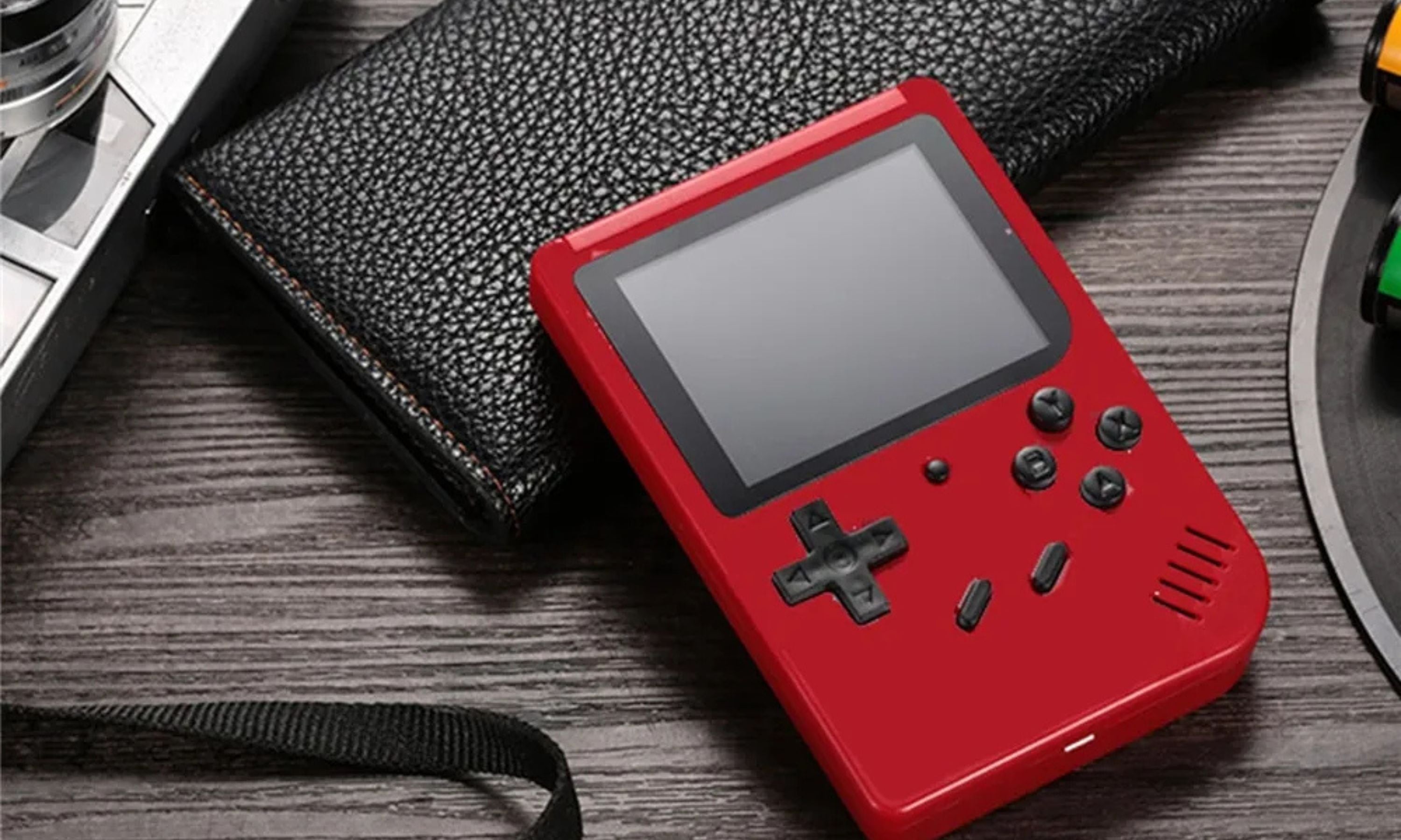 A red handheld gaming device with a D-pad, buttons, and a small screen lies on a wooden surface next to a textured black wallet. Nearby is a partial view of a camera and colorful objects.