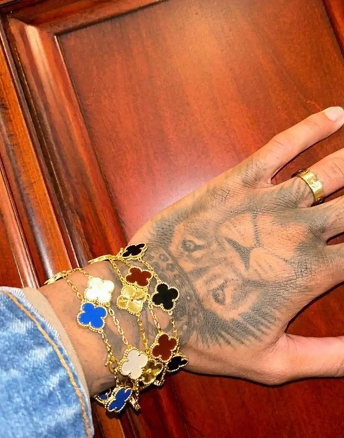 Hand with lion tattoo wearing gold chain clover bracelets in black, blue, white, and red enamel