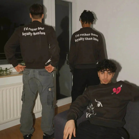 Three men indoors wearing streetwear sweatshirts, two with 'I'd rather have loyalty than love' text, one with a broken heart graphic.