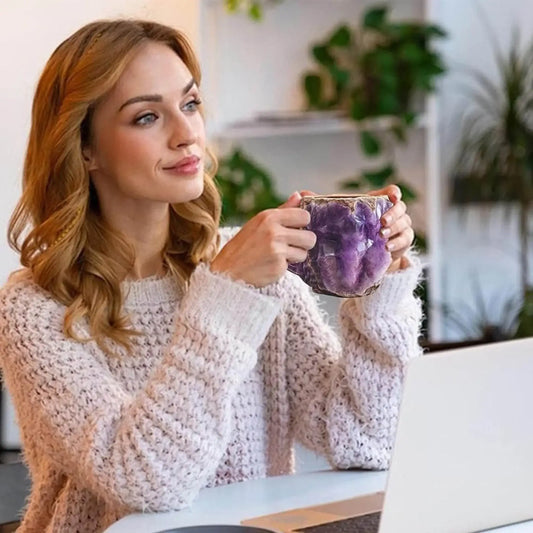 Woman in cozy sweater holding a purple crystal mug at a desk with a laptop, indoor setting