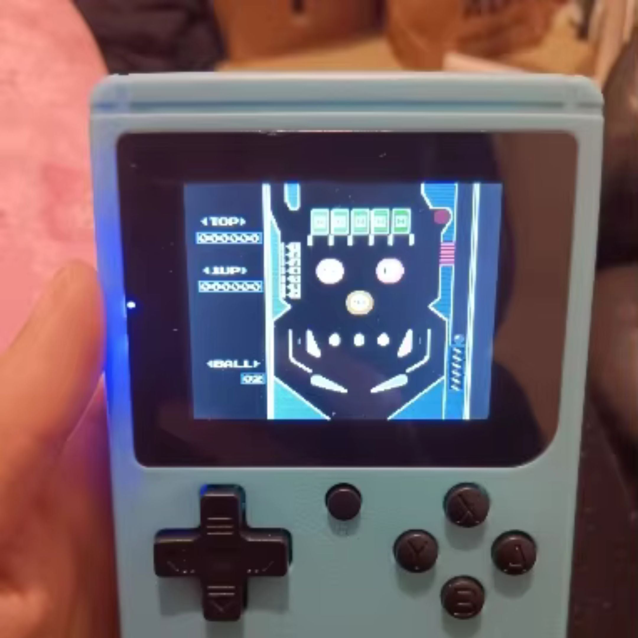 A handheld gaming device with a light blue casing displays a classic pinball game on its screen. The device has a black directional pad and four buttons. A persons hand holds it on the left side.