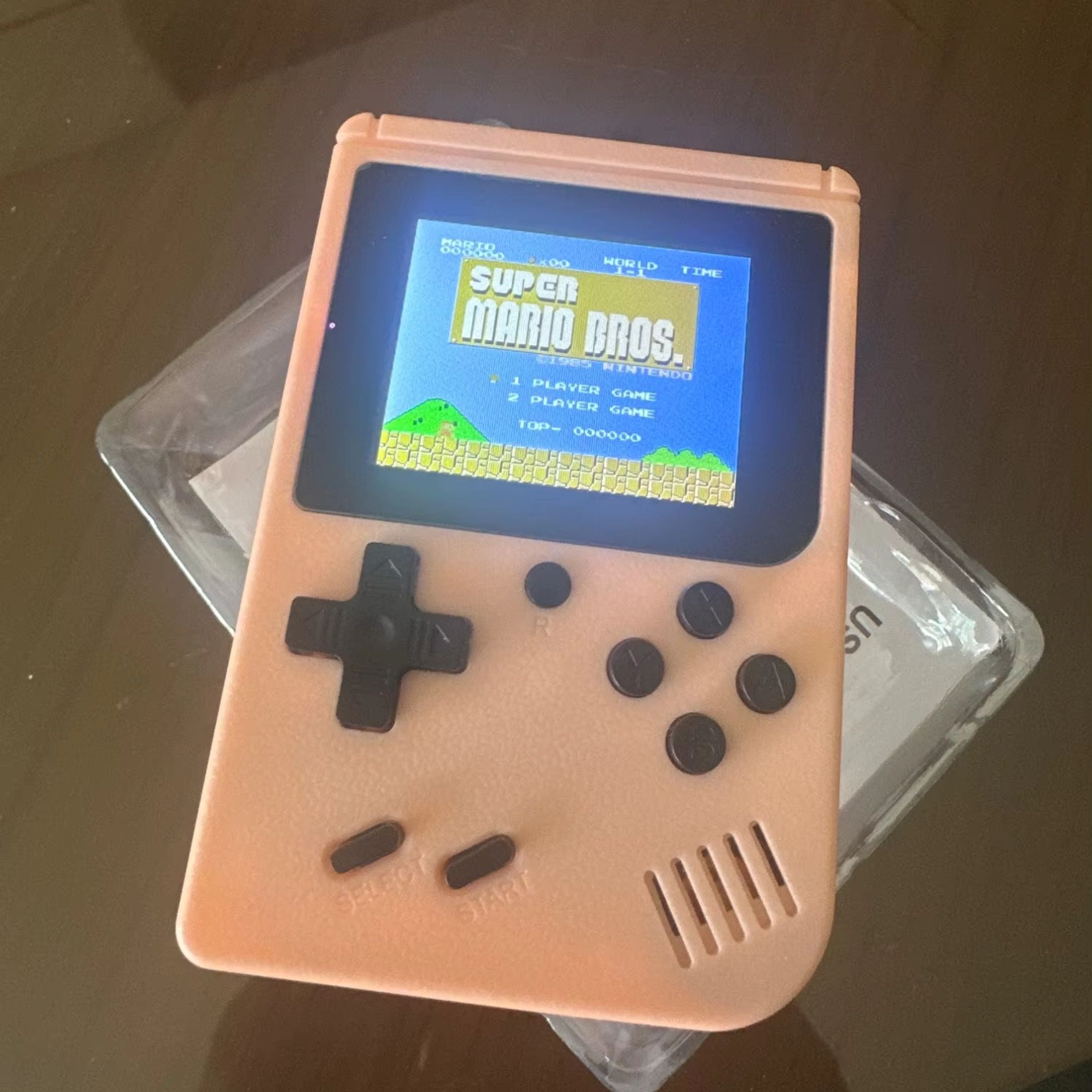 A pink handheld gaming console displays the Super Mario Bros. title screen. The console features a directional pad and several buttons. It rests on top of translucent packaging on a reflective surface.